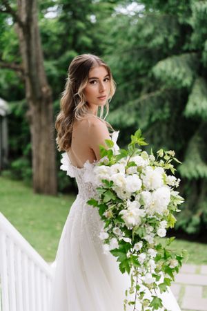 A bride in a strapless gown holds a cascading white floral bouquet on an outdoor porch.