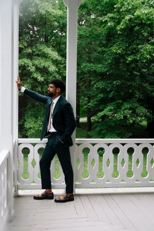 A groom in a dark suit poses casually against a white porch column with greenery in background.