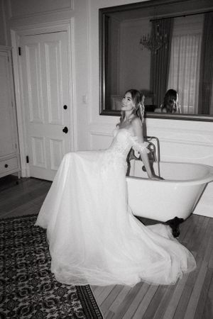 A black and white photo of a bride in a flowing gown posing near a vintage bathtub.
