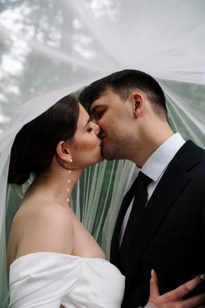 A tender kiss between newlyweds under a flowing veil with soft lighting.