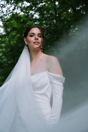 An off-shoulder bridal portrait with flowing veil and dramatic fog effect in a forest setting.