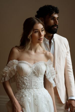A stylish wedding couple poses in white formal attire against a neutral studio background.