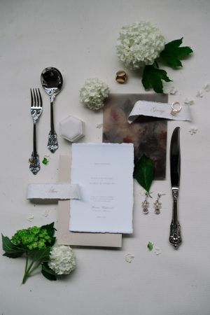Elegant wedding flatlay with white hydrangeas, vintage silverware, and invitation suite on light background.