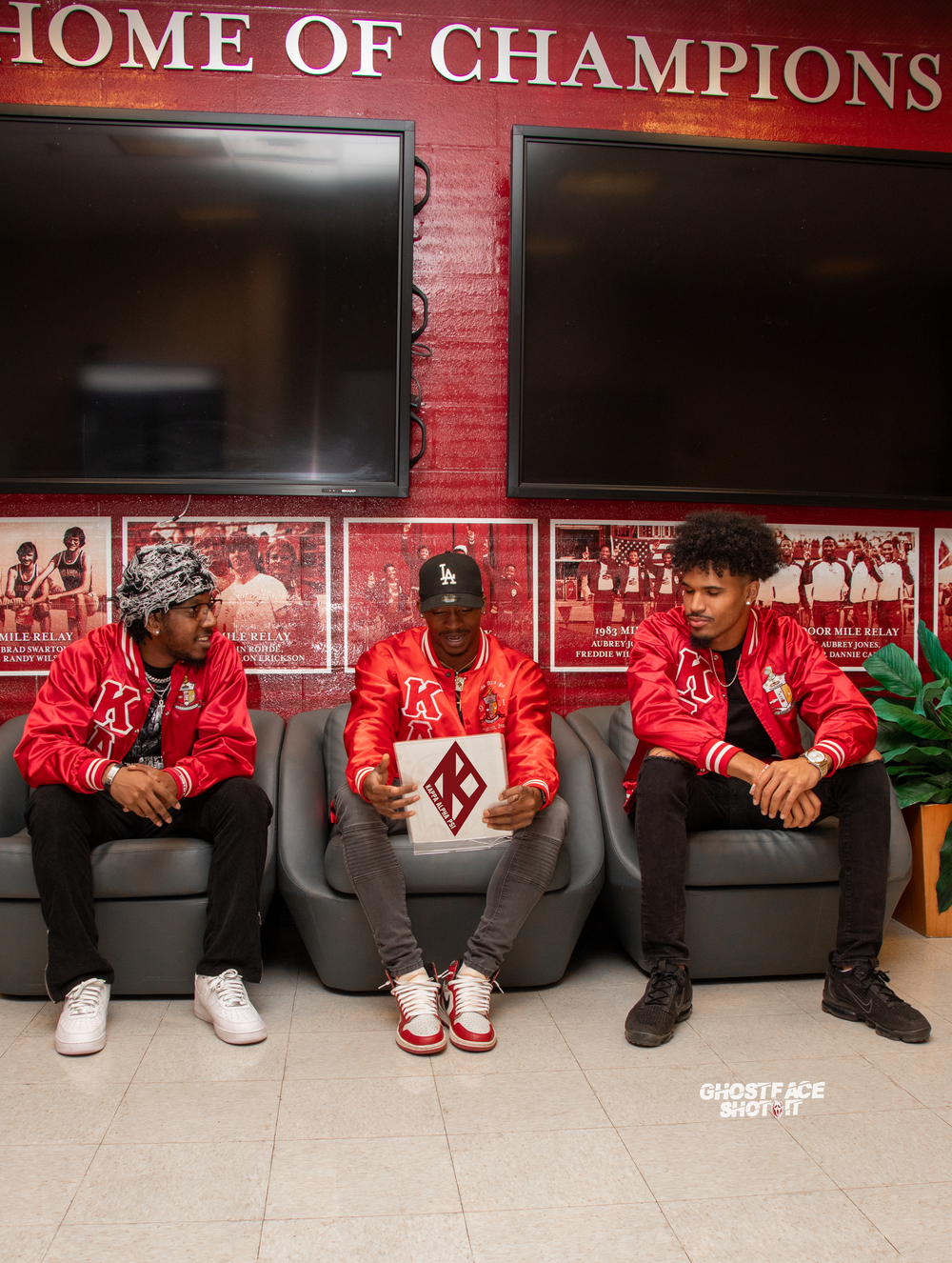 Three people in red jackets sitting beneath 'Home of Champions' sign.