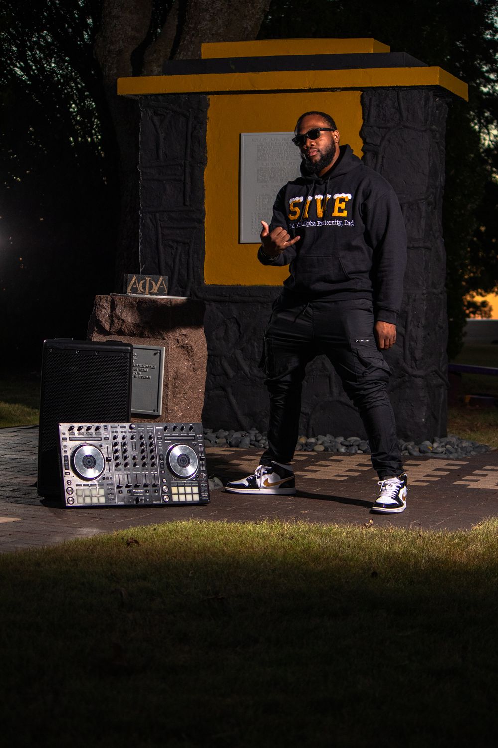 DJ with equipment poses at night against black and yellow backdrop wearing 'SOVE' hoodie.