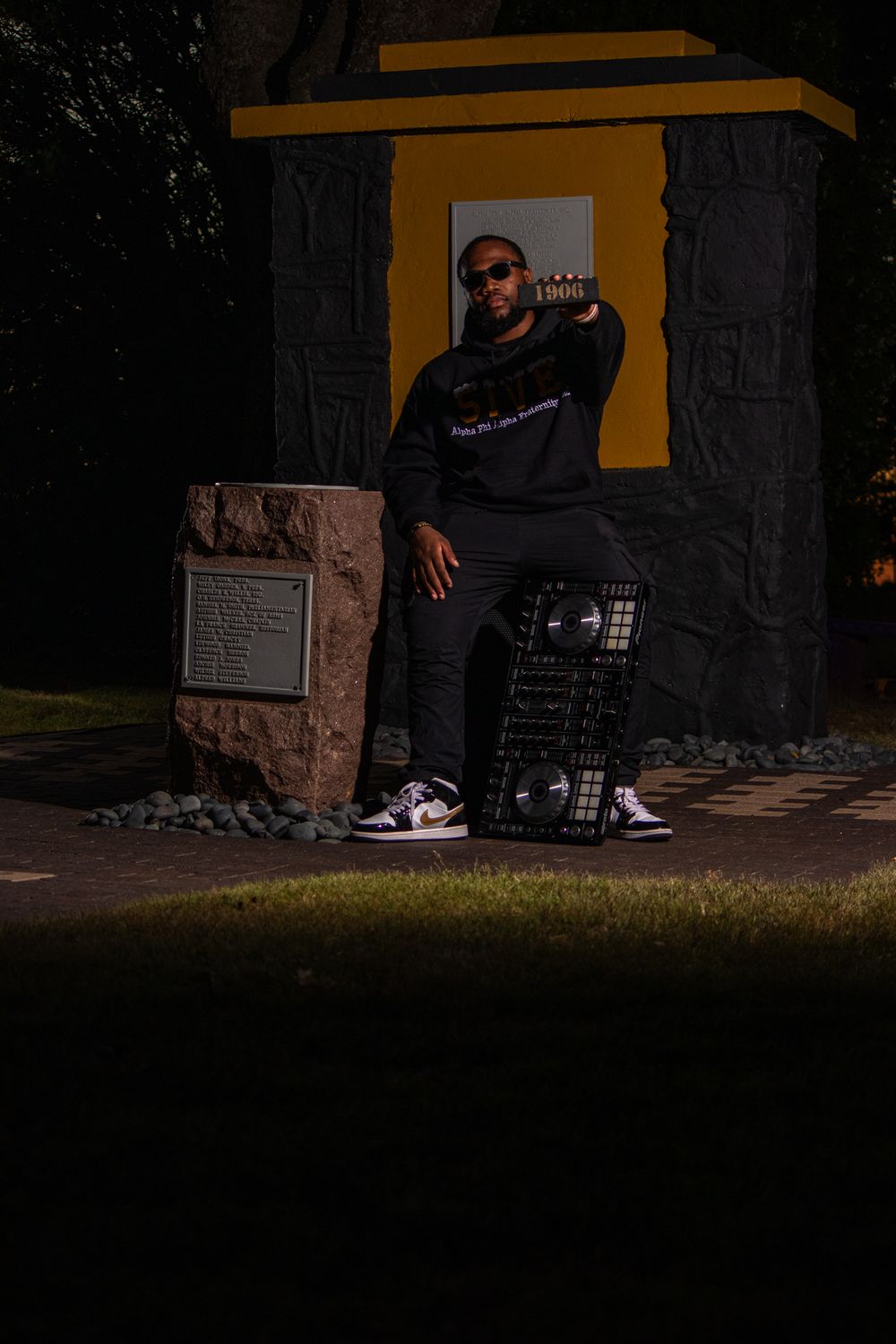Low-light portrait of DJ with equipment against urban backdrop with yellow geometric shapes.