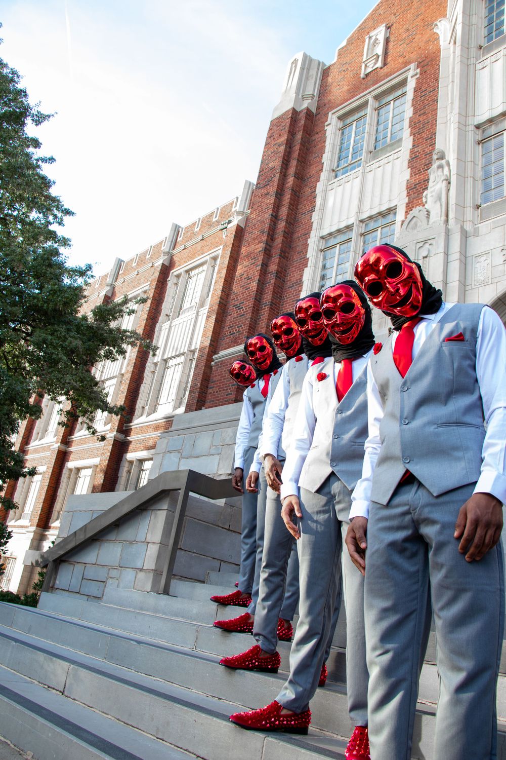 Line of people in grey suits and red masks stand on stone steps of brick building.