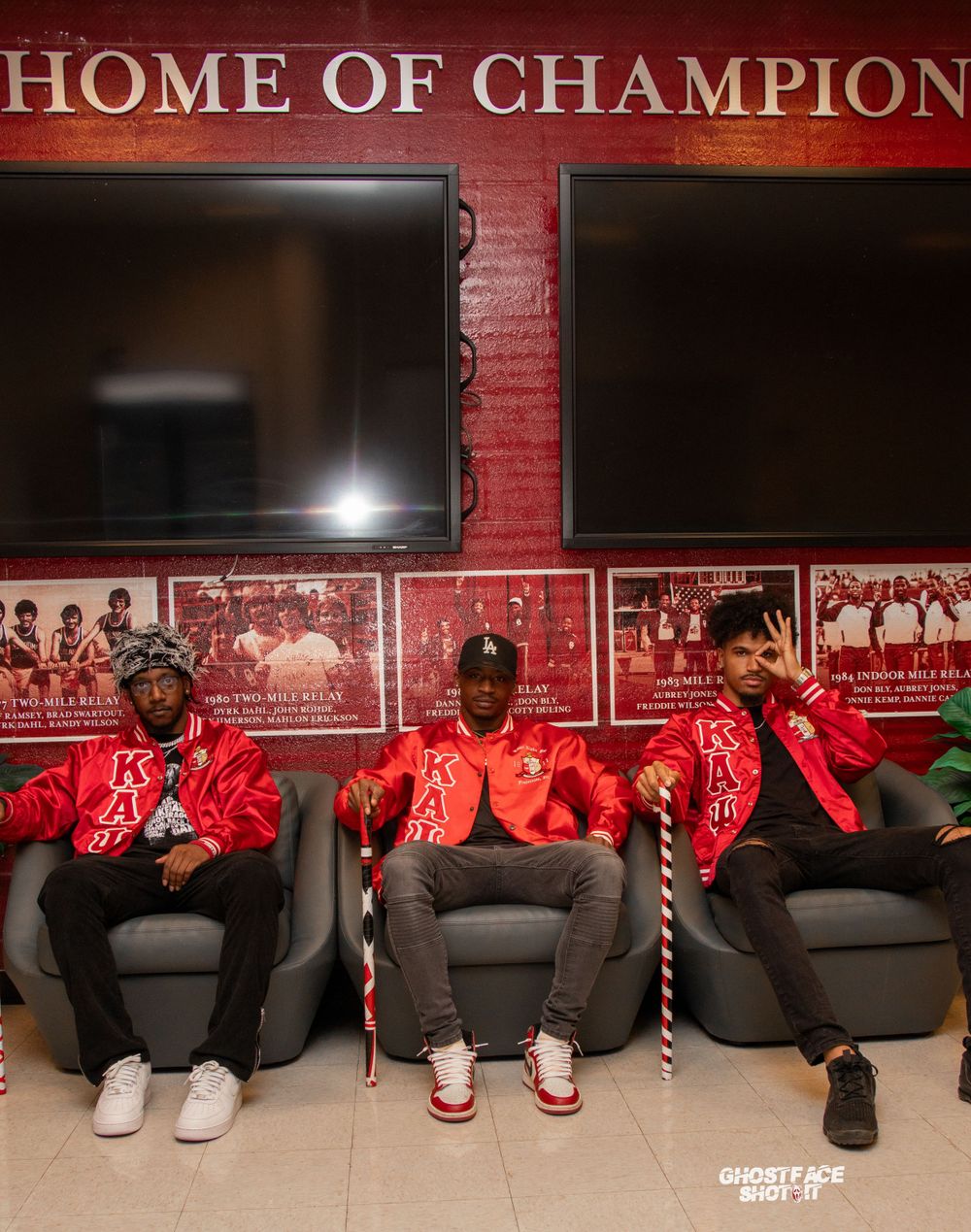 Three people in red jackets sit on chairs beneath 'Home of Champions' sign.