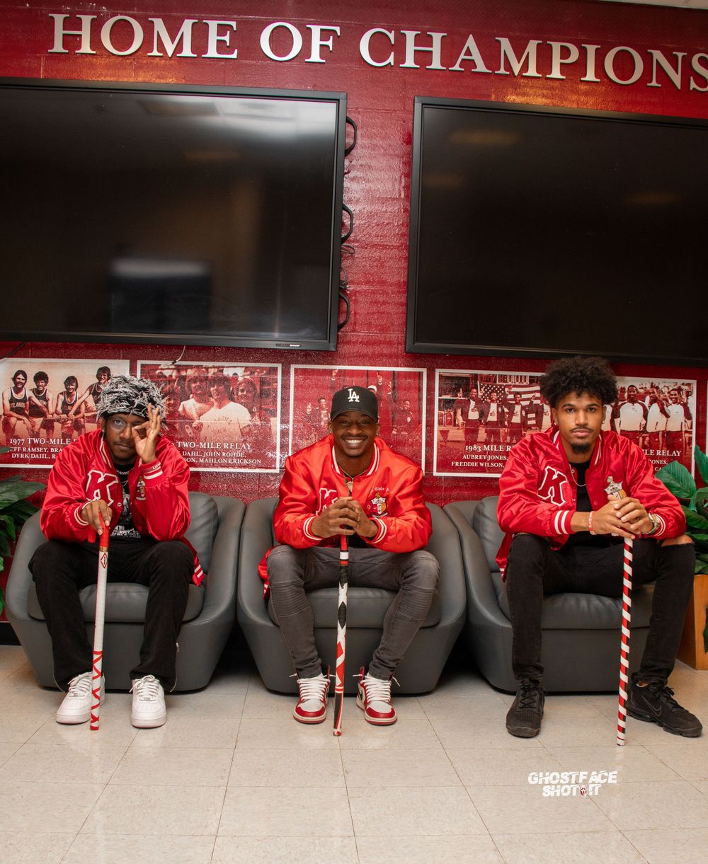 Three individuals seated on chairs beneath a 'Home of Champions' sign wearing red jackets.