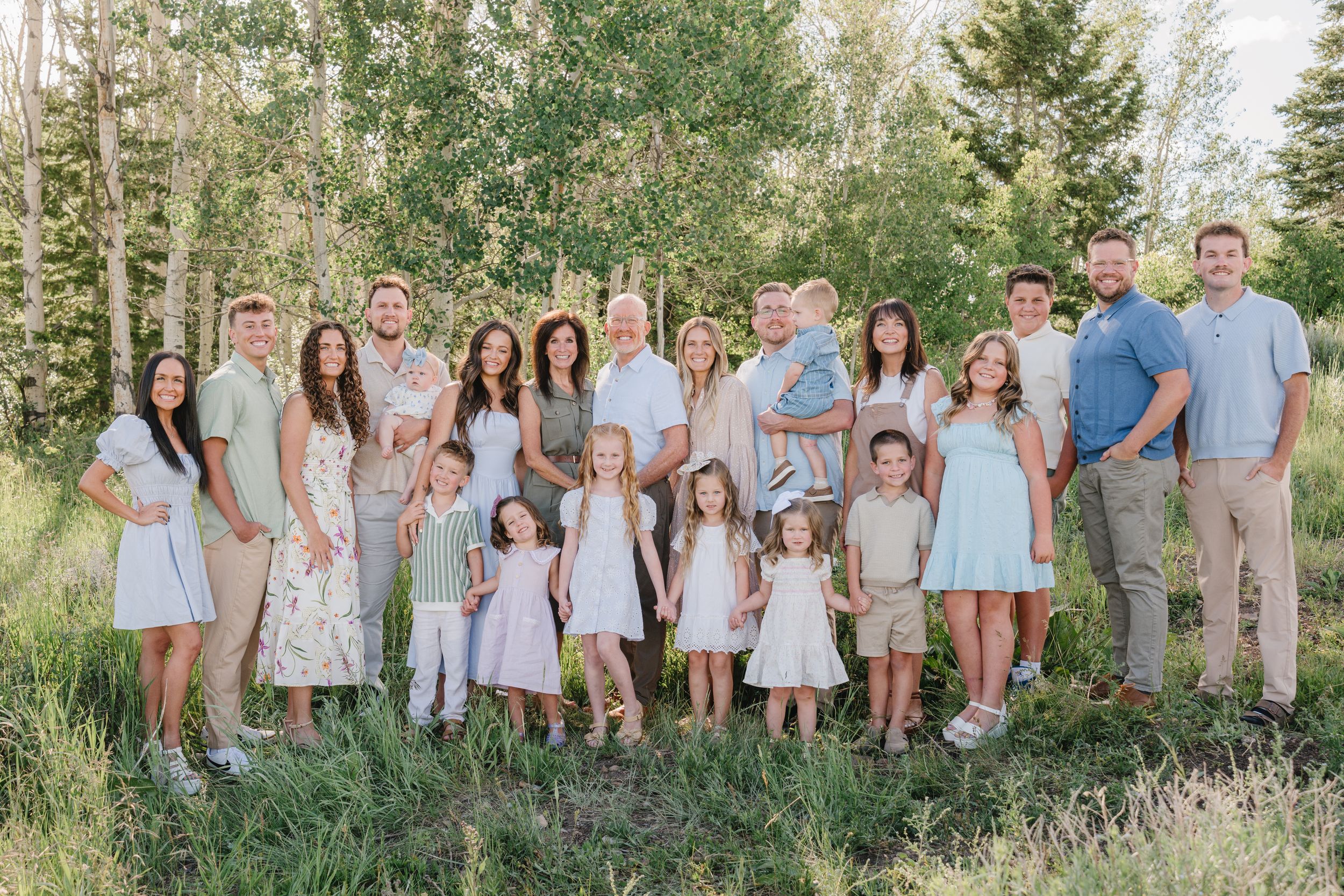 Large family group poses together on green lawn with lush trees and foliage in background during outdoor photo session.