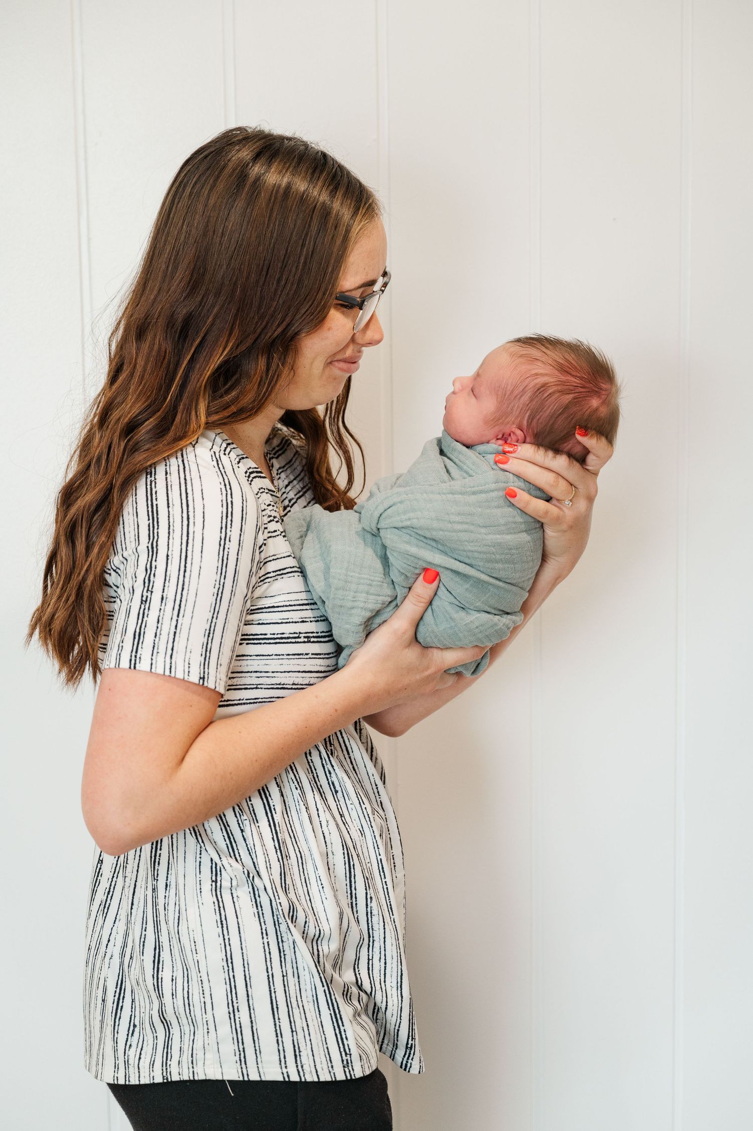 A tender moment between a caregiver in a striped dress holding a swaddled newborn baby against a white wall.