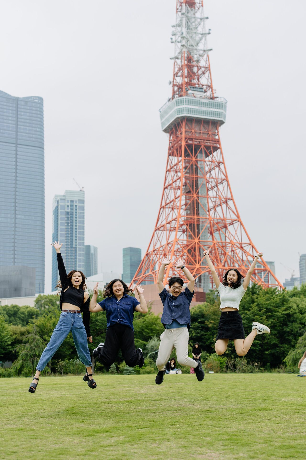 Friends jump joyfully in front of Tokyo Tower with modern buildings in the background.