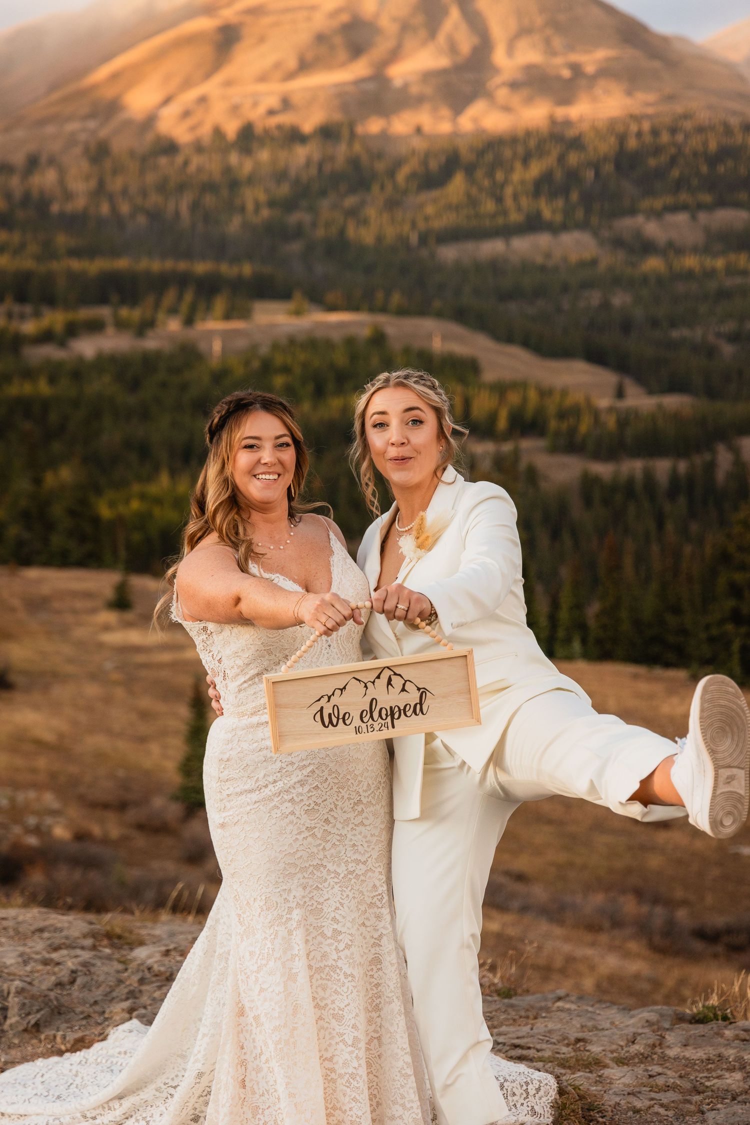 Couple in wedding attire pose together against a mountain landscape during golden hour with 'We eloped' sign.