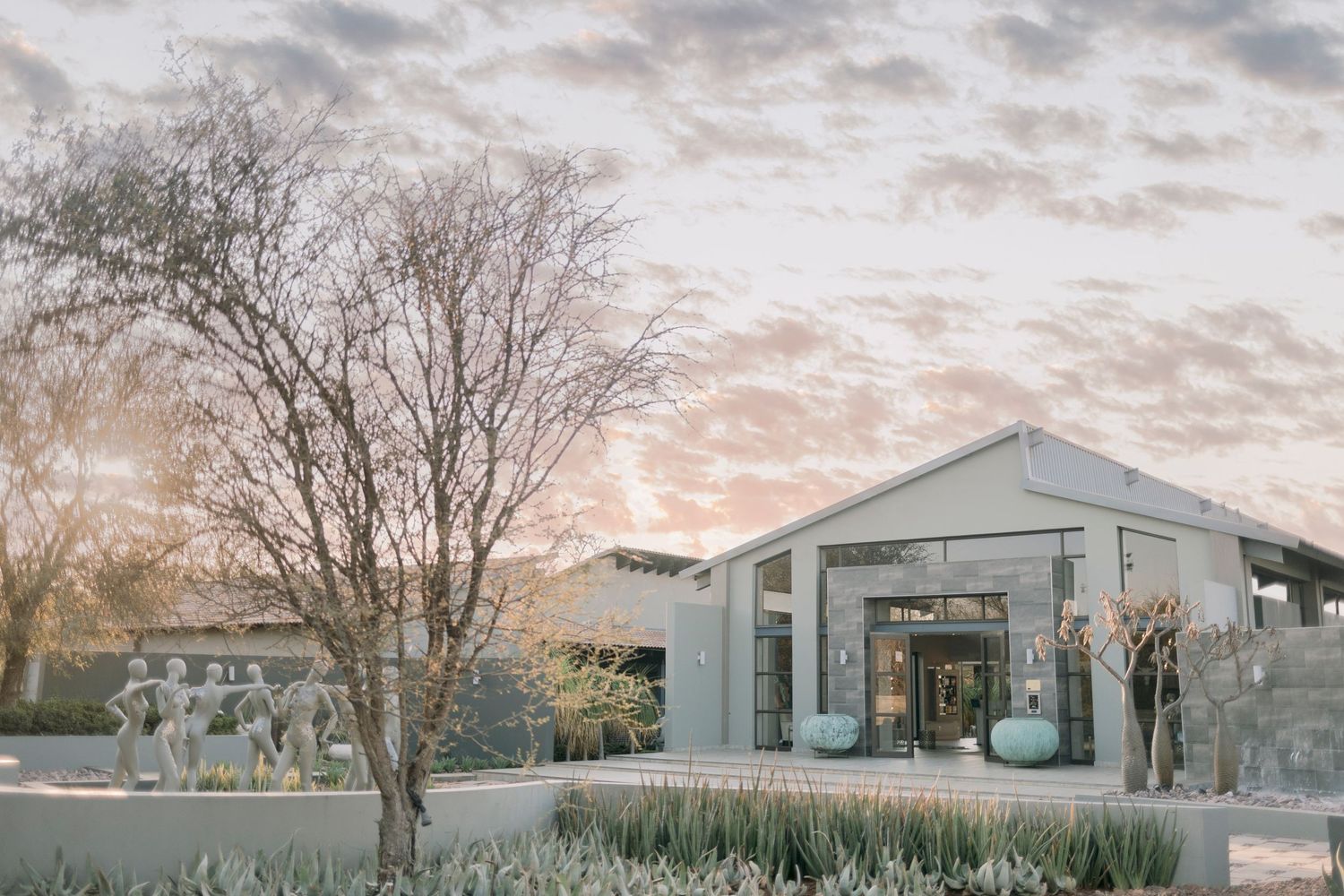 Modern luxury lodge exterior surrounded by desert plants at a soft pastel sunset.