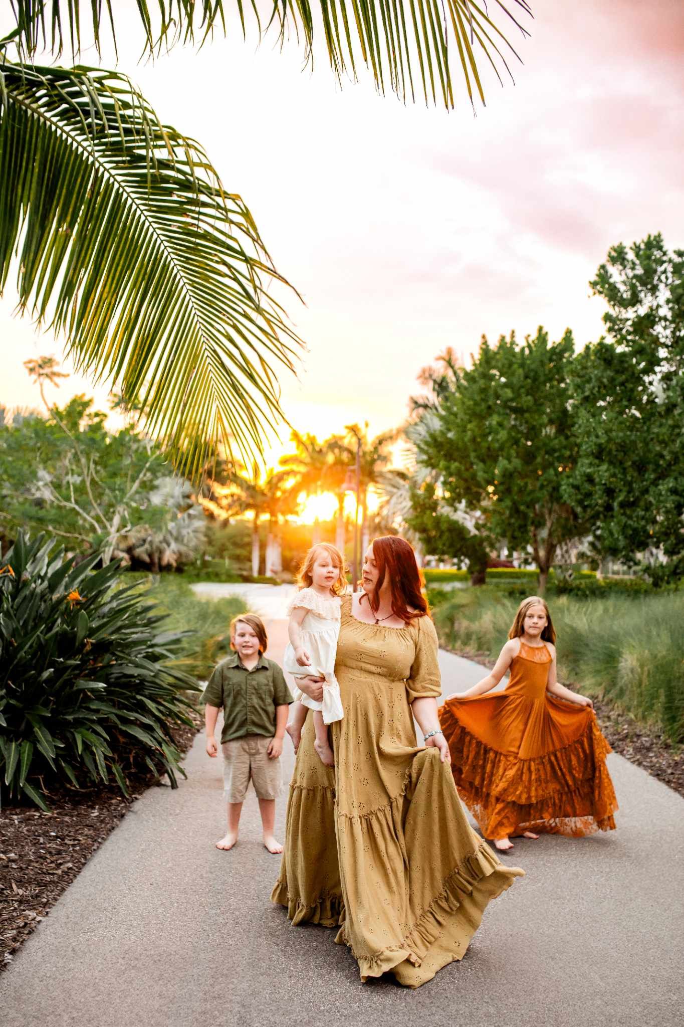 Group in coordinated earth-tone dresses twirls on palm-lined path at sunset.