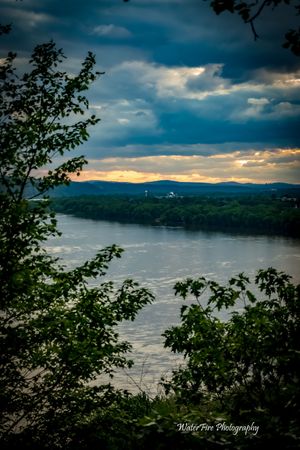 Dramatic sunset view of a river valley with mountains in the distance through tree branches and moody cloudy skies.