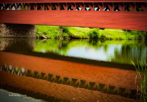 Red covered bridge spans across water with decorative triangular trim along its length.