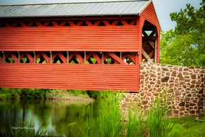 Side view of red covered bridge with stone foundation reflecting in peaceful water.