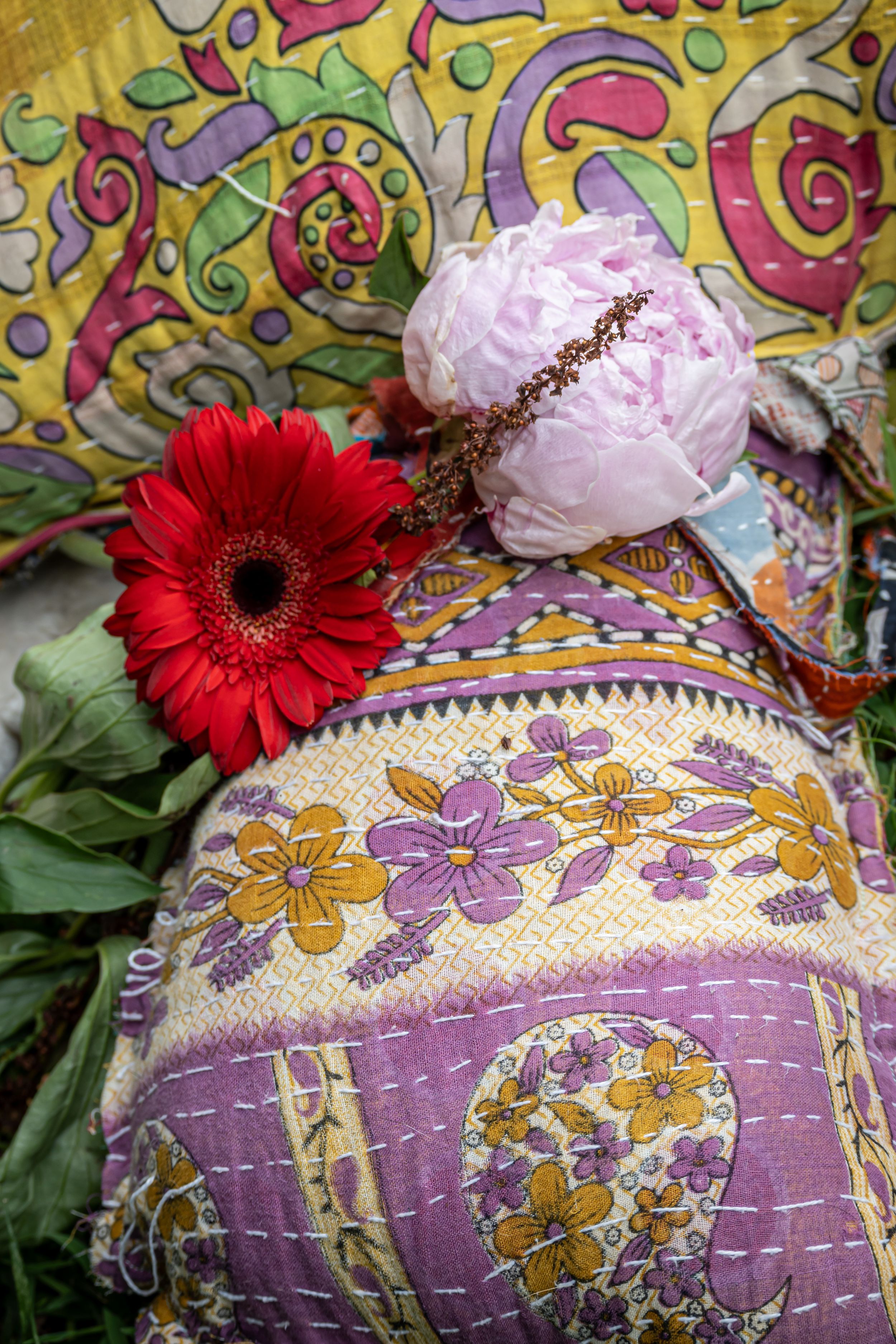 Vibrant red gerbera daisy and pink peony against colorful floral patterned pillow with purple and yellow embroidery.
