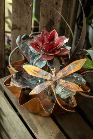 A decorative copper and metal flower sculpture with a red bloom and dragonfly detail sits on a rustic wooden surface.