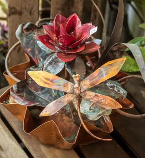 Handcrafted metal and glass floral art pieces with copper petals and stems catch light on a wooden surface.