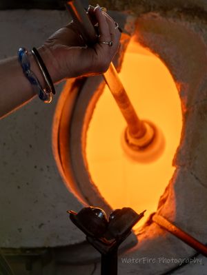 Molten glass being poured in circular motion from a metal ladle, glowing bright orange against dark background.