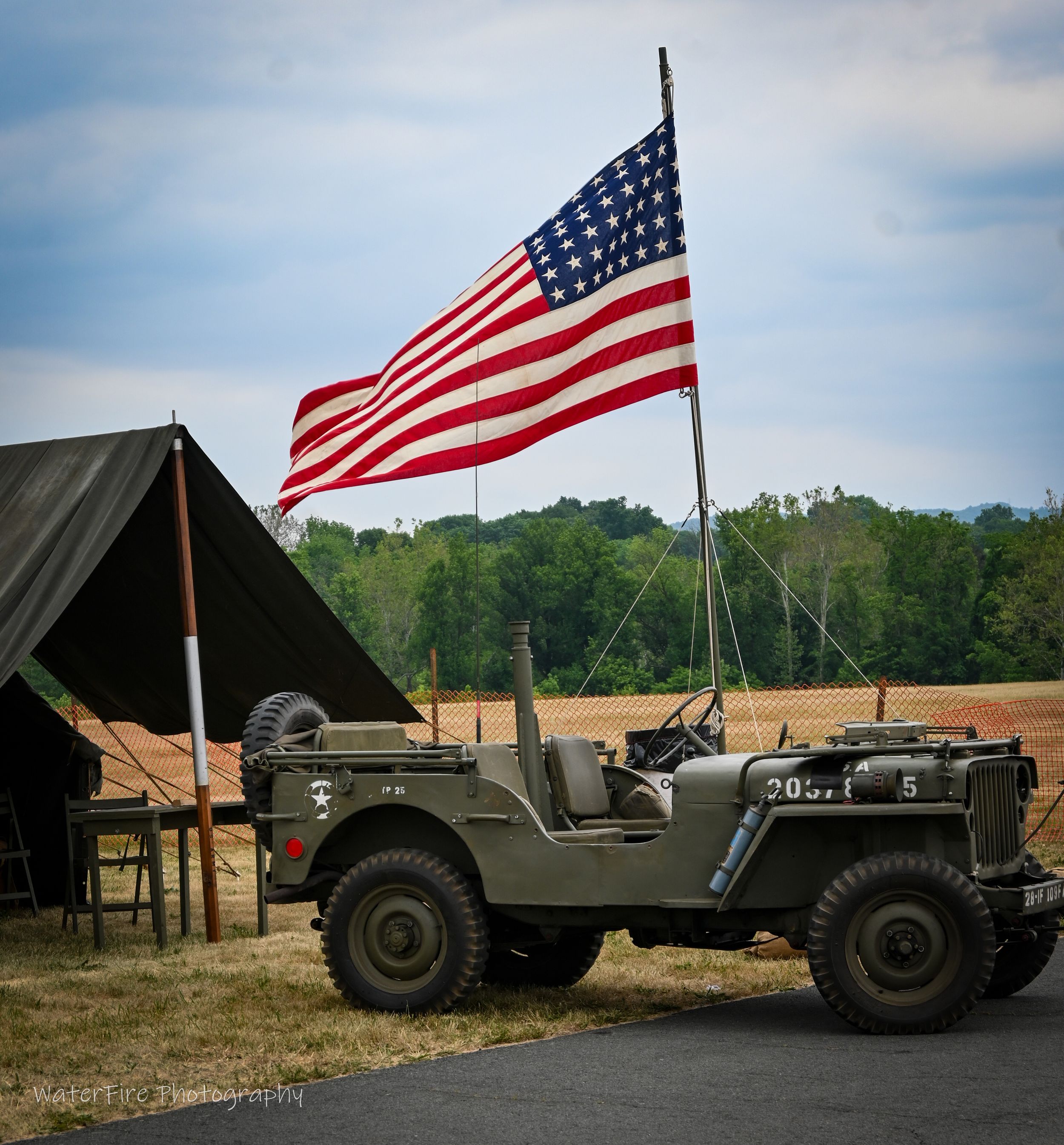 A vintage military Jeep with American flag displayed next to an olive green tent in a rural outdoor setting.