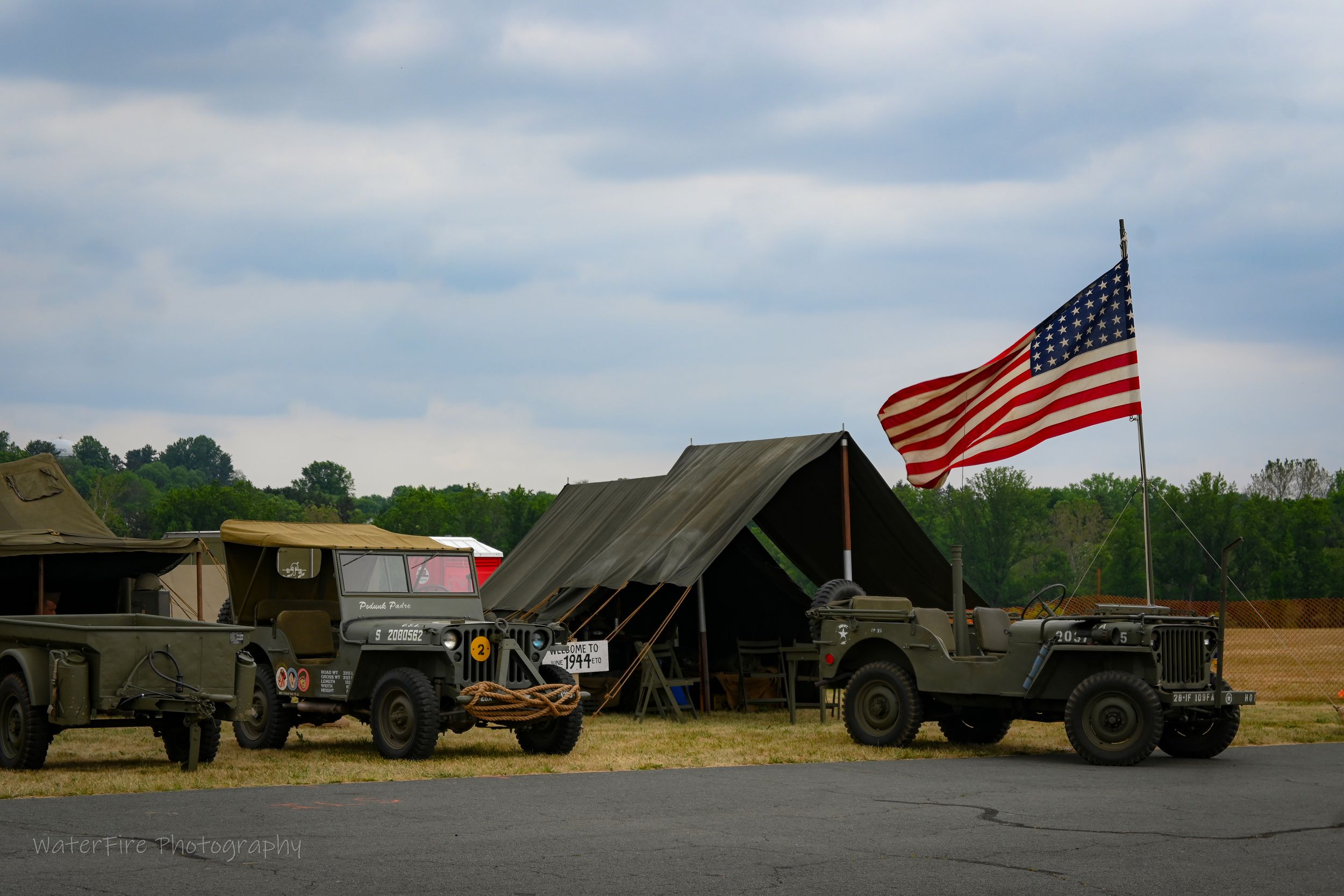 Military vehicles and tent setup with American flag flying at outdoor encampment during historical reenactment event.