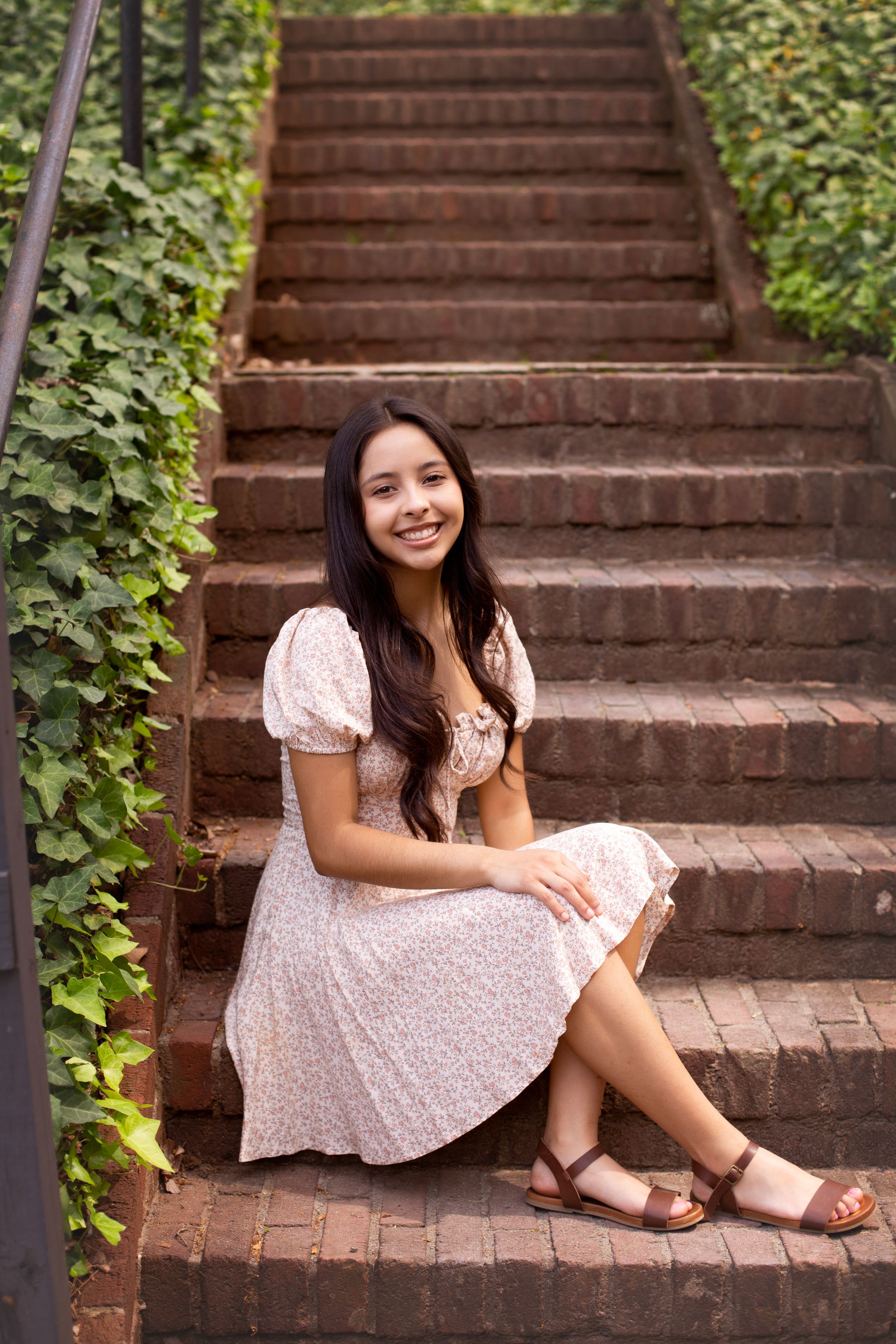 Senior girl in a floral dress, sitting on a brick staircase surrounded by greenery.
