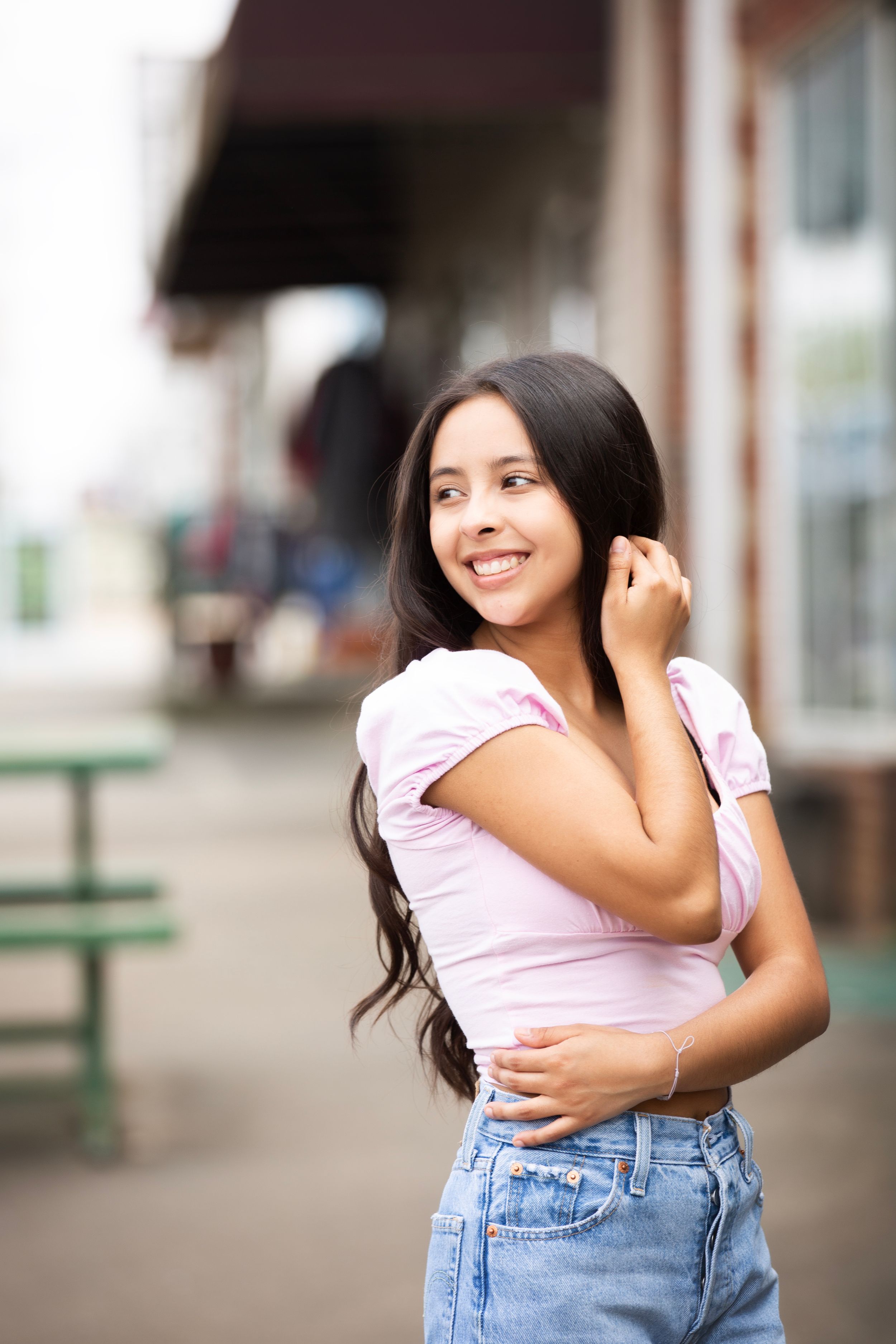 Senior girl standing on the side walk in a small town. She's wearing a pink top and jean pants.