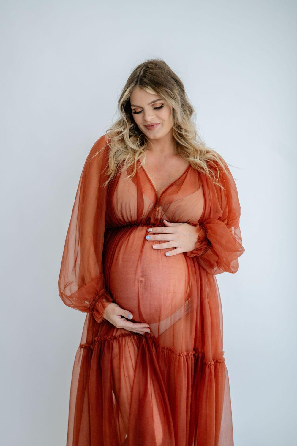 Maternity studio portrait featuring rust-colored sheer gown against white background.