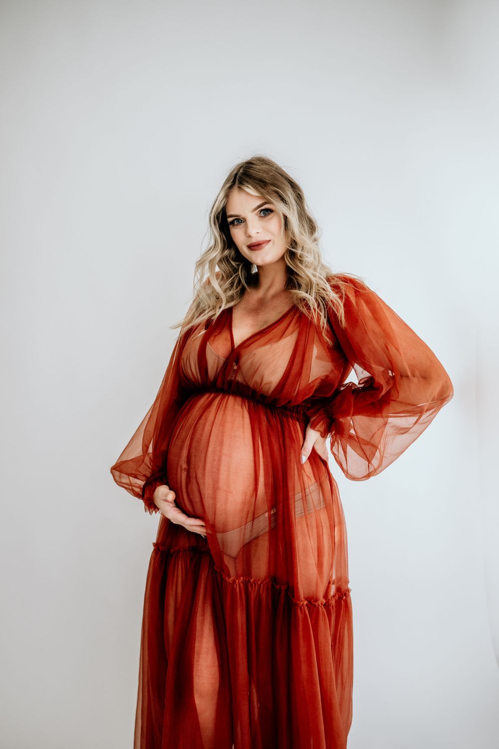 Elegant maternity pose in flowing rust-colored dress in studio setting.
