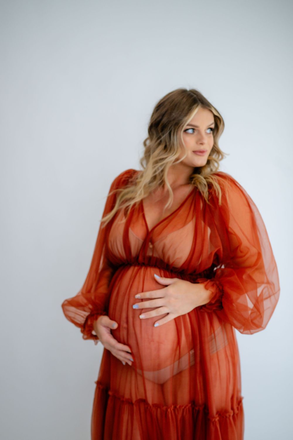 Close-up studio portrait featuring a sheer rust-colored maternity gown with billowing sleeves.