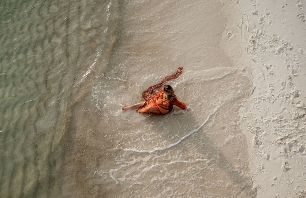 Aerial view of a person in orange dress lying on beach near water's edge.