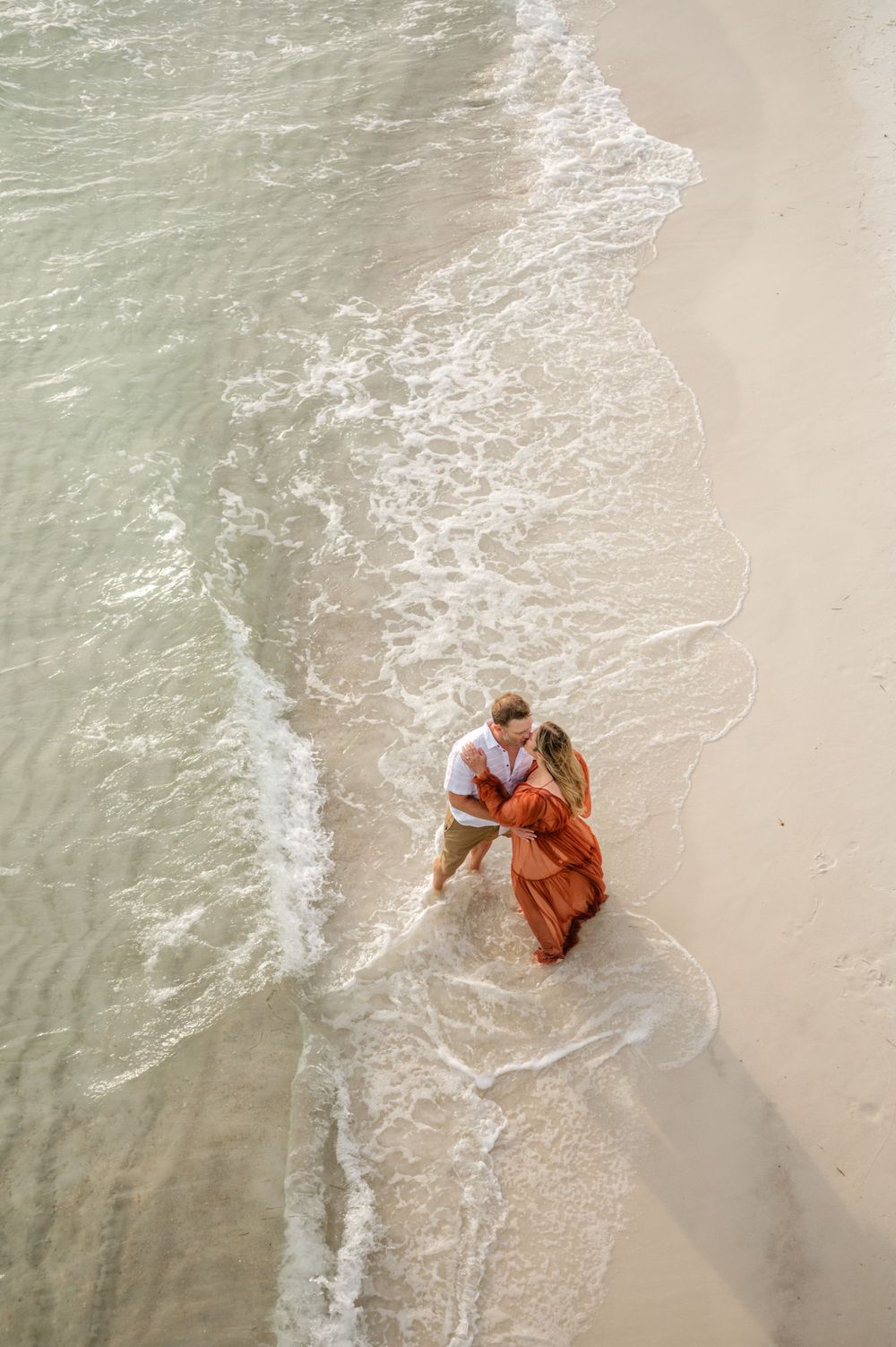 Aerial beach photo showing couple embracing at water's edge with waves rolling in.