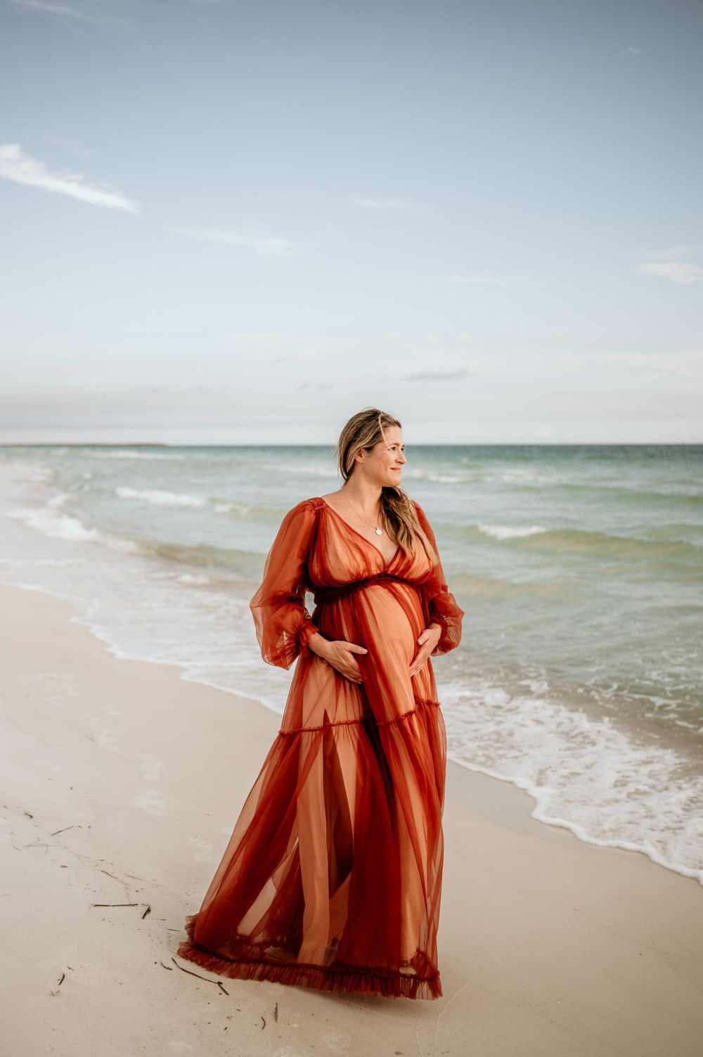 Seaside maternity portrait featuring flowing red dress against ocean waves.