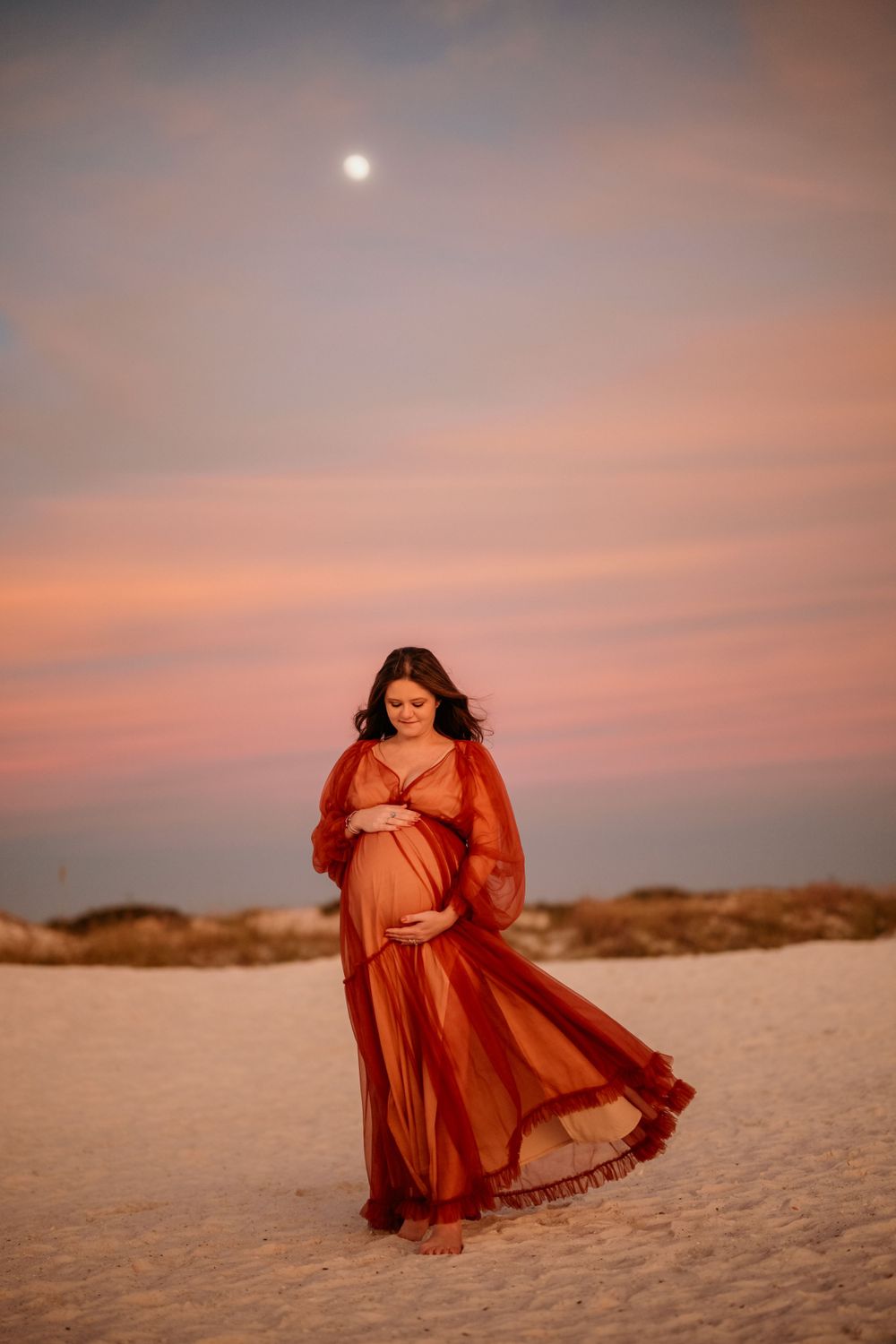Dramatic beach maternity photo with full moon and flowing red dress.