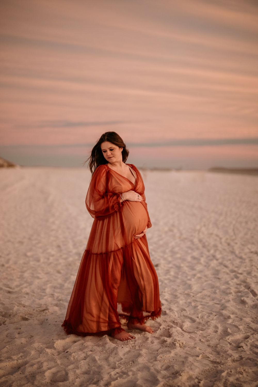 Expectant mother in a flowing red dress poses on sandy beach at sunset.