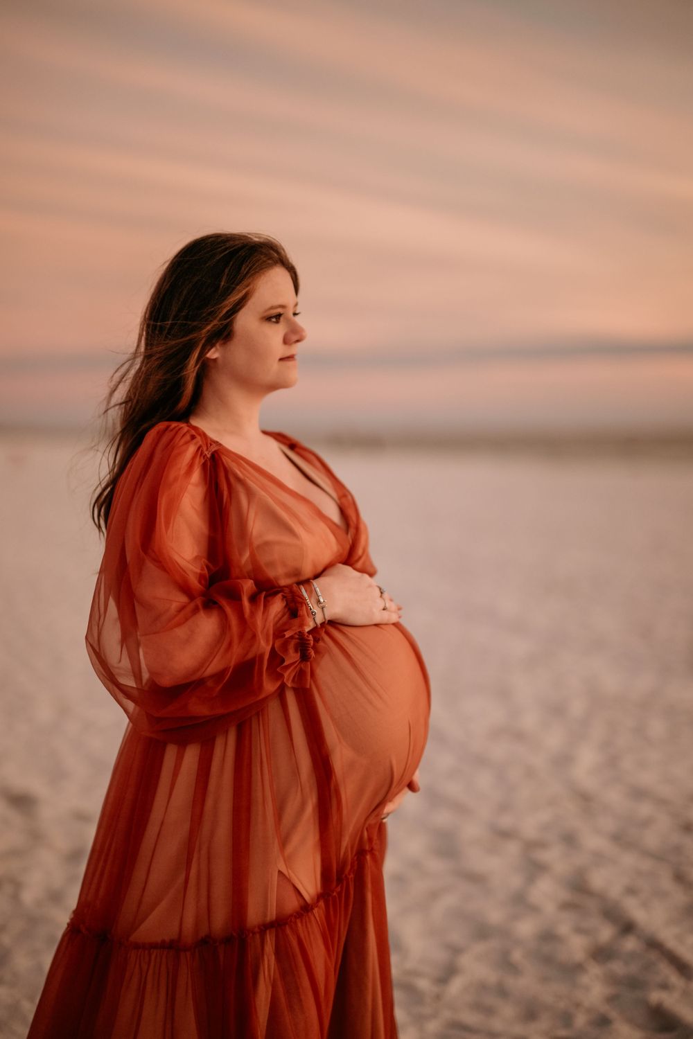 Sunset maternity photo on a beach with a flowy rust-colored dress.