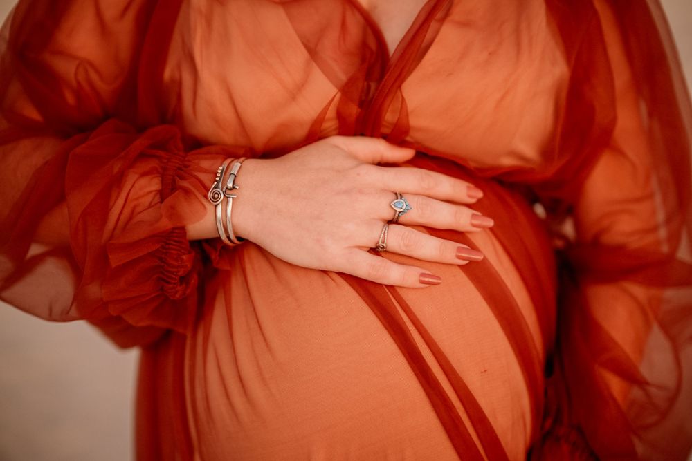 Close-up detail shot of jewelry and hands cradling a pregnant belly through sheer fabric.