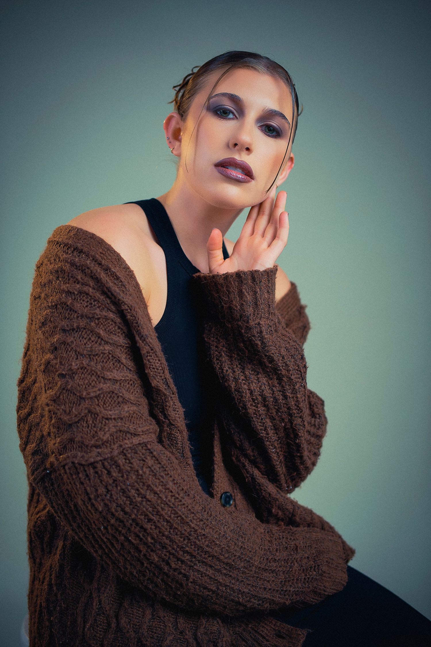 Fashion portrait of model in brown knit cardigan and dark top against teal backdrop with dramatic makeup.