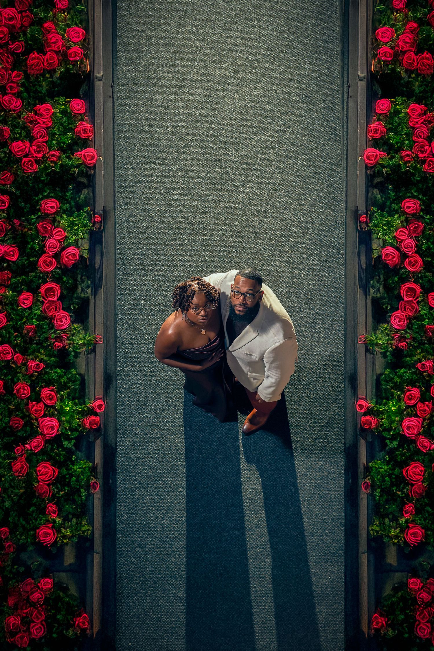Aerial view of a couple standing on green carpet between vibrant red rose bushes creating symmetrical borders.