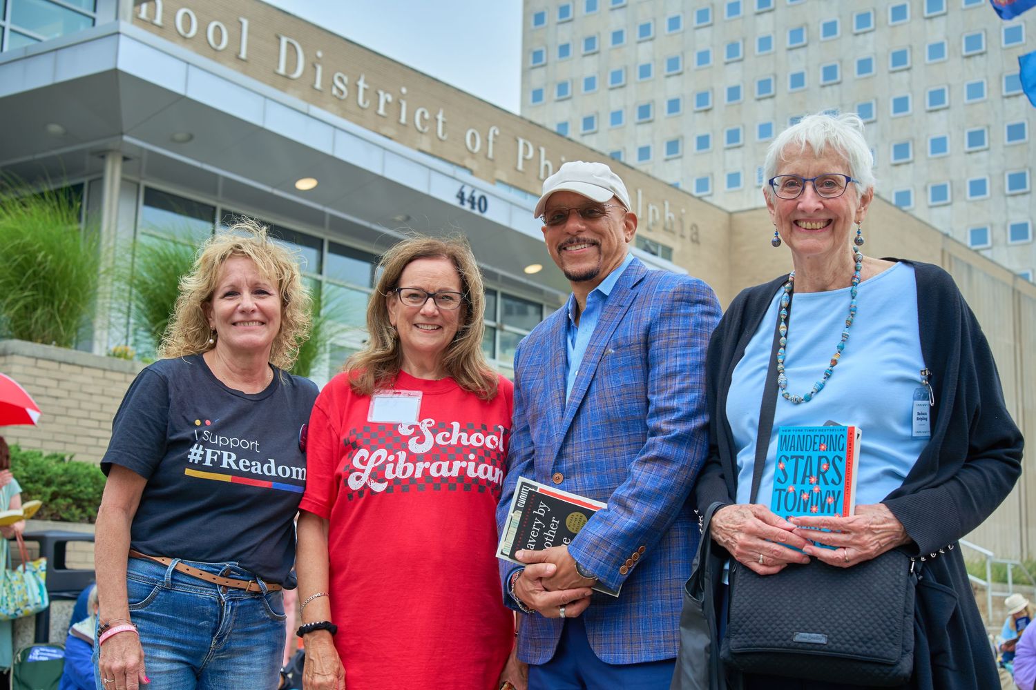 Four people stand together outside a school district building holding books while smiling at the camera.