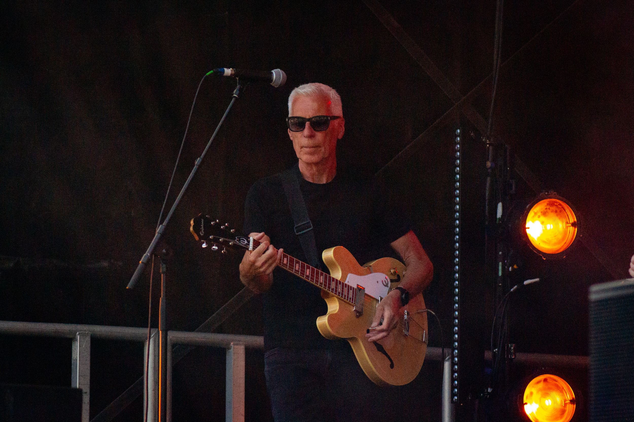 Guitarist in black attire plays a vintage hollow body guitar on stage with orange spotlights.