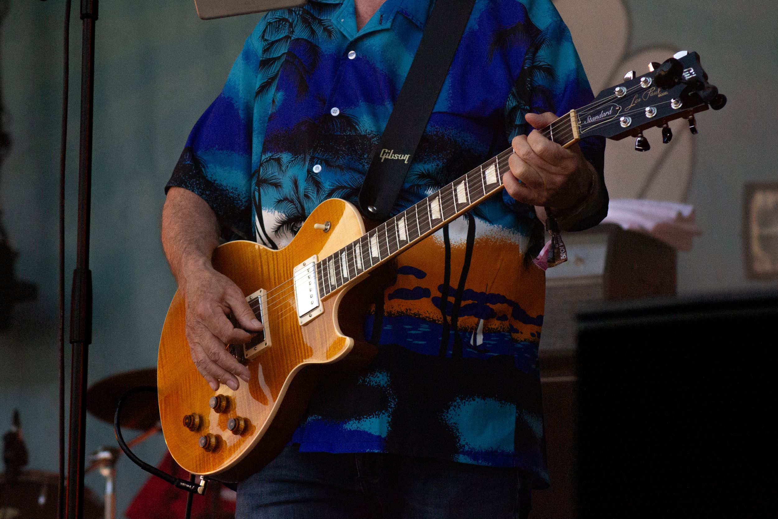 Close-up of hands playing sunburst Les Paul guitar against blue backdrop on stage.