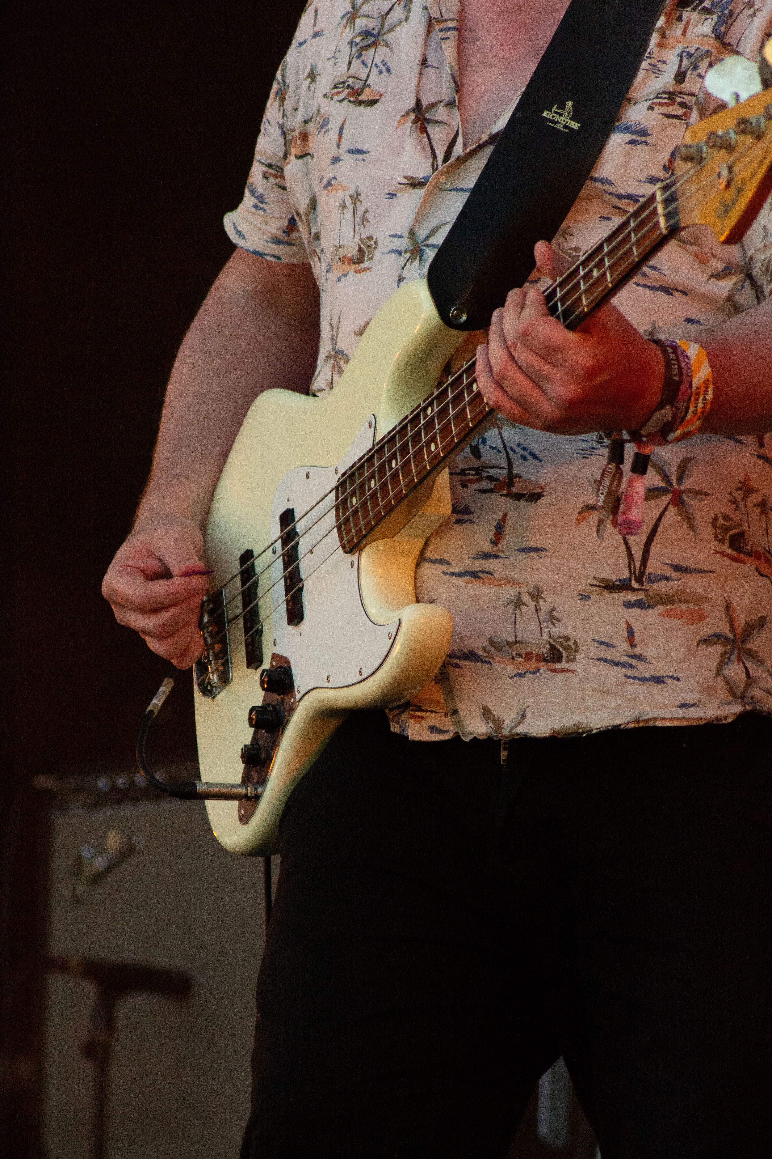 Musician plays cream colored Fender bass guitar while wearing tropical print shirt on stage.