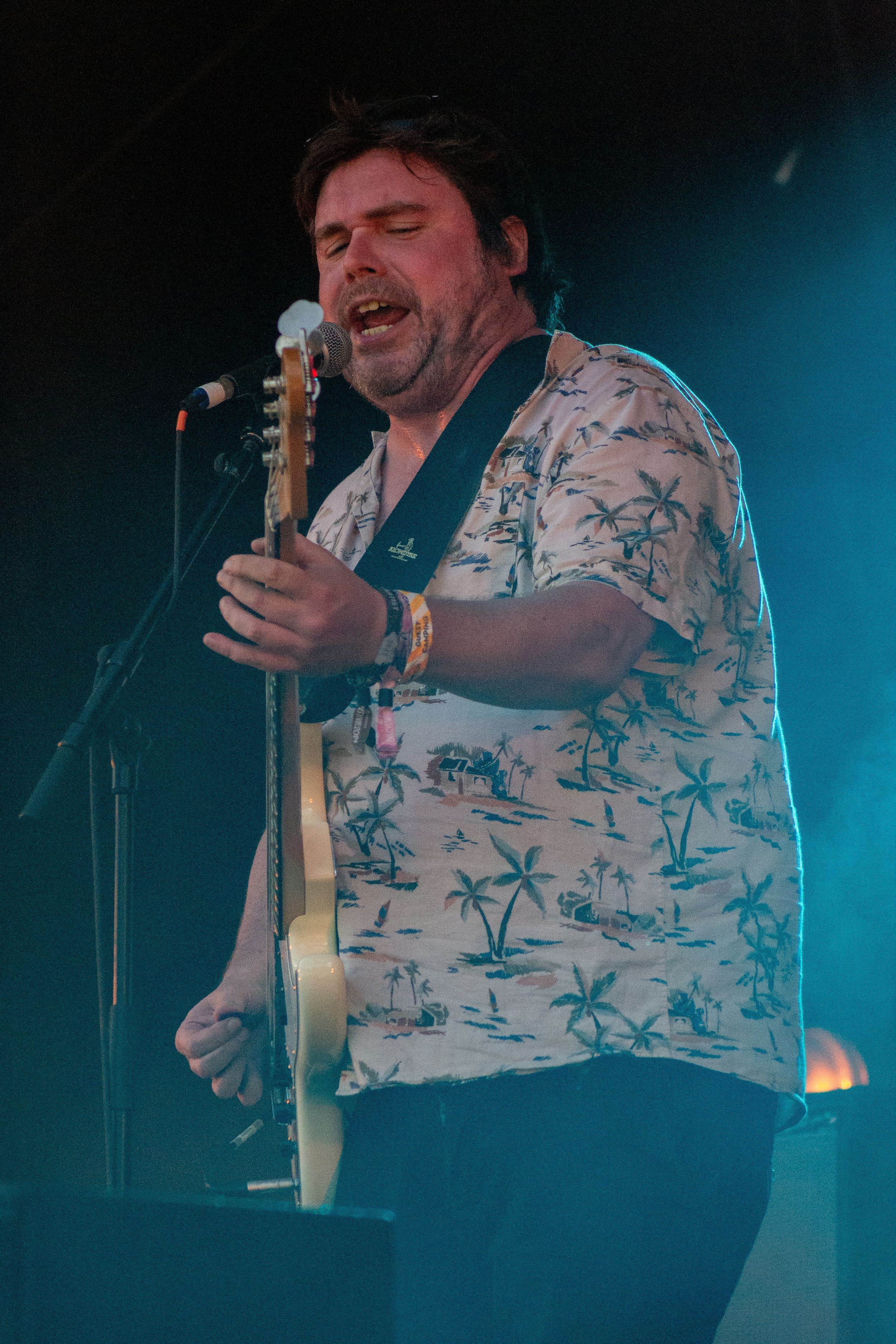 Performer in palm tree print shirt plays guitar passionately against blue stage lighting.