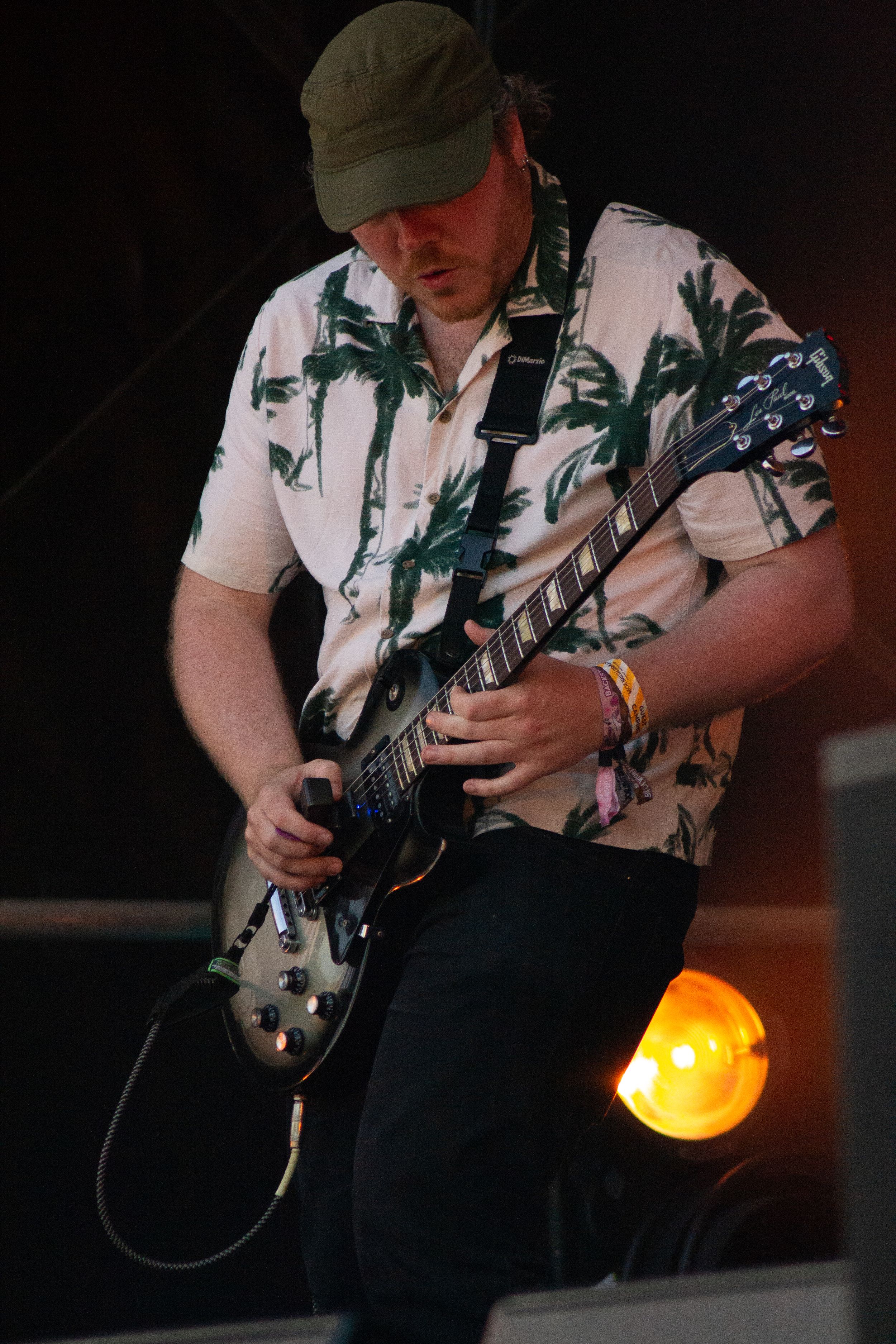 Guitarist wearing cap and tropical shirt performs solo on dark stage with amber lighting.