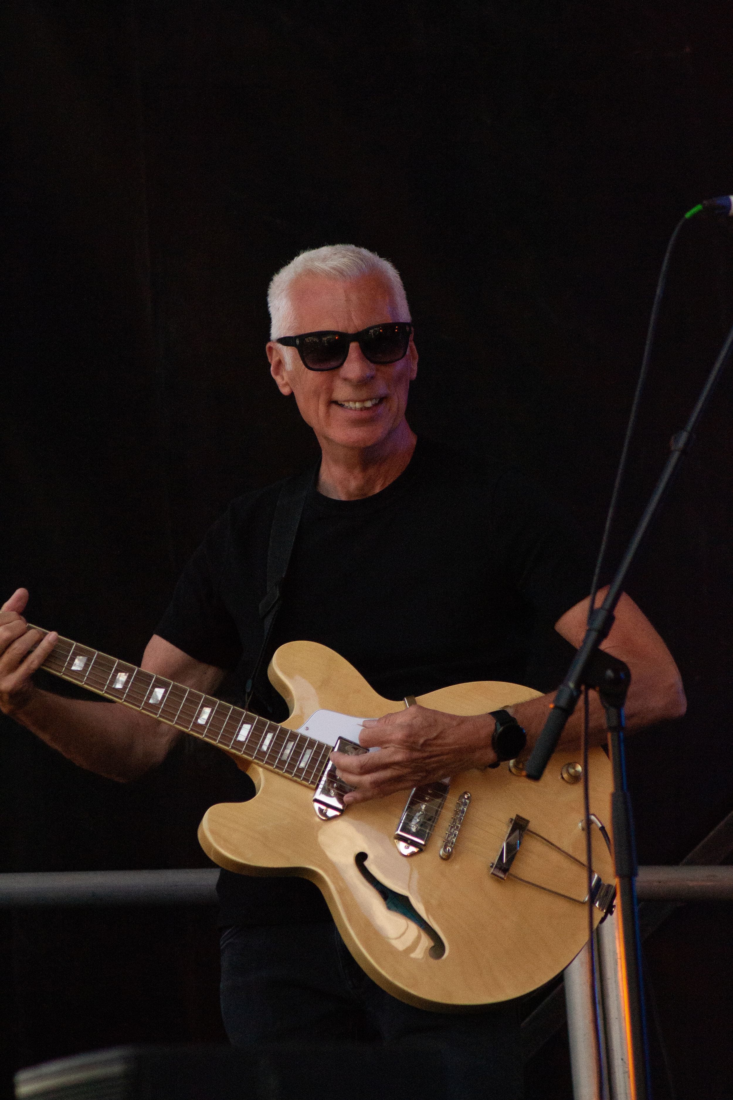 Performer with natural hollow body guitar smiles on dimly lit concert stage.