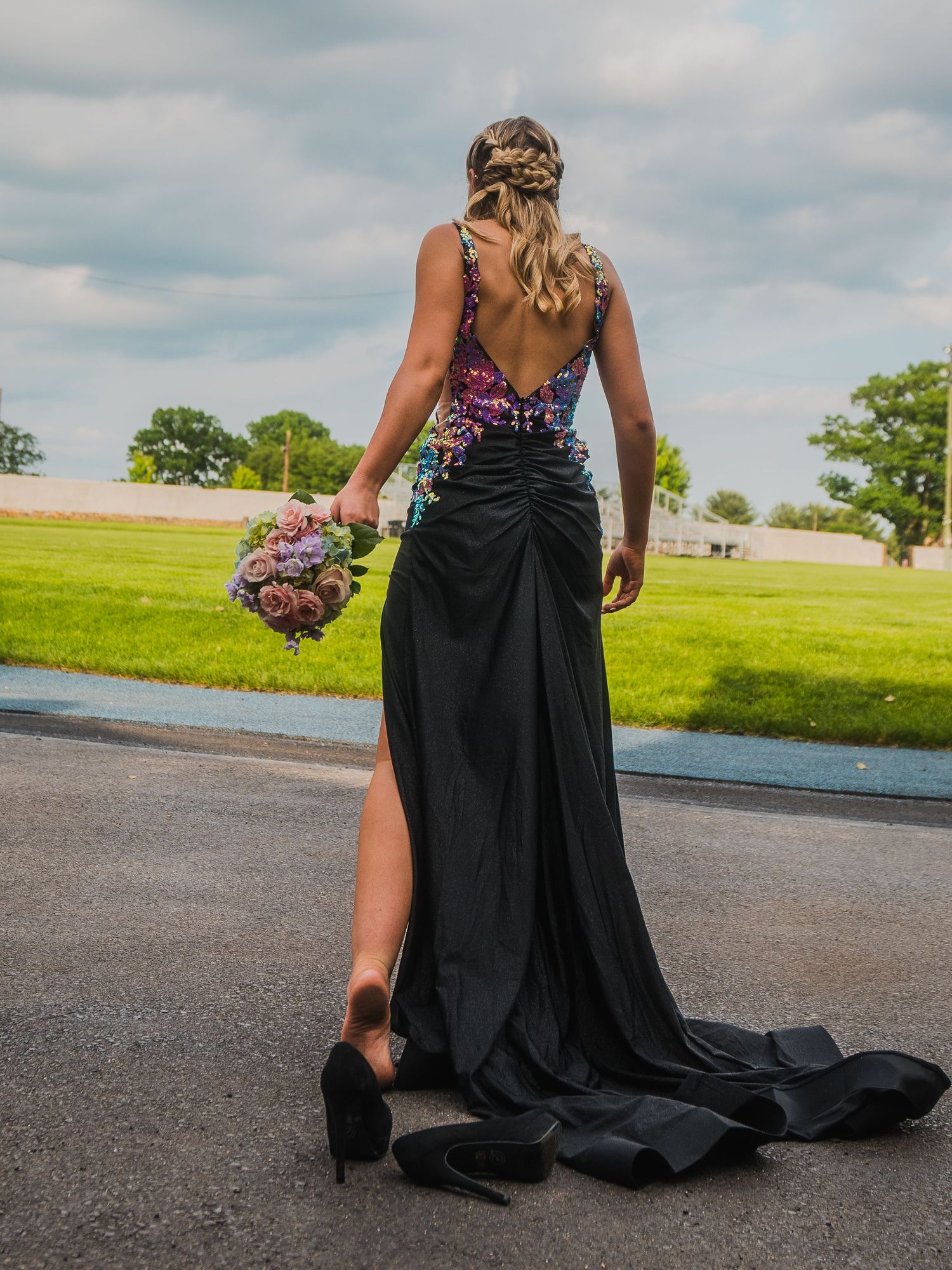Person in elegant black backless gown holding colorful bouquet stands on road against green field and cloudy sky.